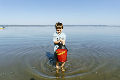 Boy carrying bucket while standing in sea against clear blue sky during sunny day