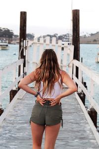 Rear view of woman with hands behind back standing on pier by sea in city