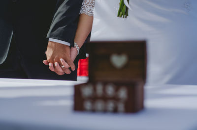 Midsection of bride and groom holding hands with jewelry box on table