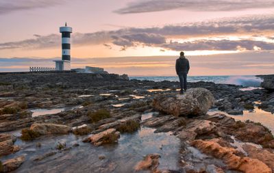 Man standing on rock by sea against sky during sunset