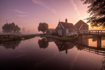 Reflection of houses and trees in lake against sky during sunset