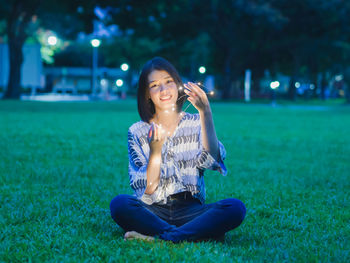 Young woman holding illuminated string lights while sitting on grassy field