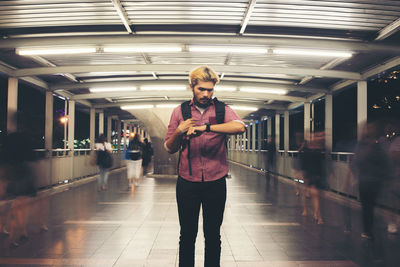 Young man looking at wristwatch while standing on footbridge at night