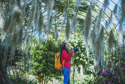 Side view of woman holding camera while standing in forest