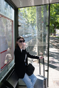 A business woman in sunglasses drinks water at a tram stop in the city, dressed
