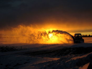 Tractor splashing snow against orange sky during sunset