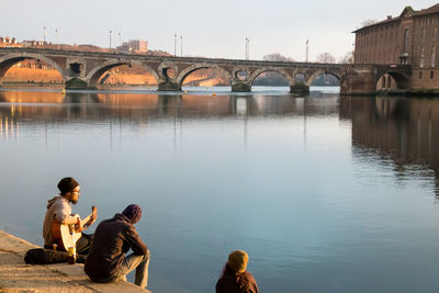 People sitting on arch bridge over river against sky