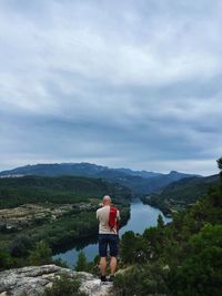 Rear view of man looking at view while standing on rock against cloudy sky