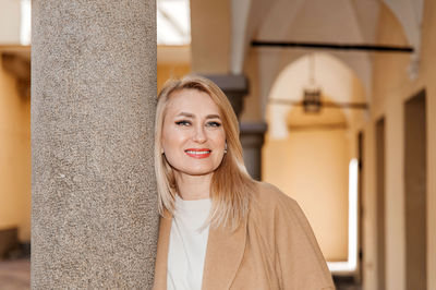 Blonde woman smiles gently leaning on a textured column in an airy corridor