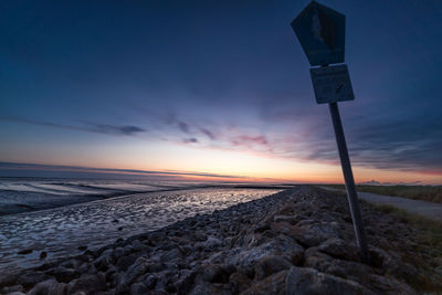 Road sign on beach against sky during sunset