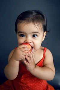 Baby girl in red tank top eating ripe red apple on black background in studio