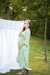 Beautiful pregnant woman with big belly stands near clothesline with bed sheet, young female takes