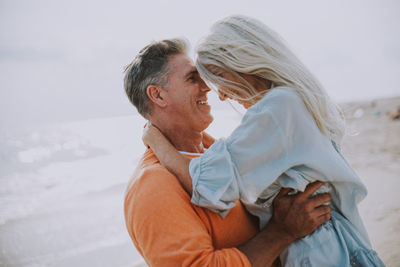 Side view of happy couple embracing at beach against sky