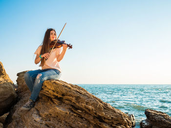 Young woman playing violin while sitting on rock by sea against sky