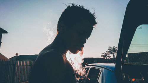 Side view of young man smoking while standing against clear sky during sunset