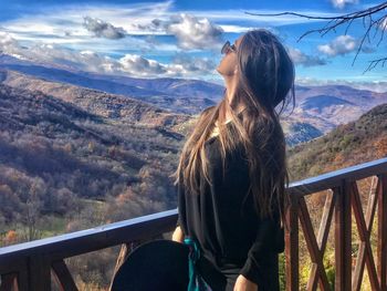 Young woman wearing sunglasses while standing by railing against sky