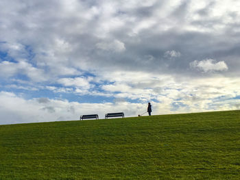 Low angle view of man with dog by benches on grassy hill against cloudy sky