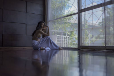 Full length side view portrait of woman sitting on floor by window at home