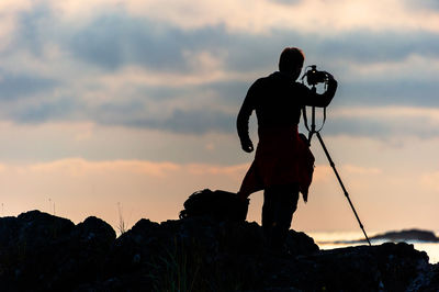 Silhouette man photographing on rock against sky during sunset