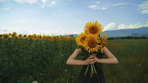 Young woman holding sunflowers while standing in farm against sky