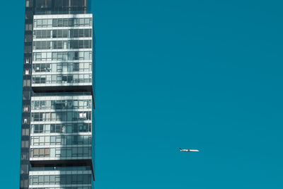 Low angle view of airplane flying by modern building in clear blue sky