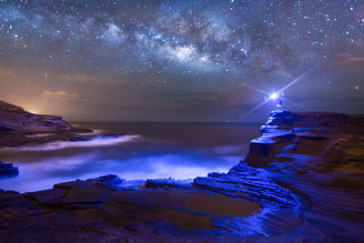 Distant view of man holding illuminated flashlight on rock at shore against star field