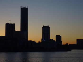 Silhouette buildings by sea against sky during sunset