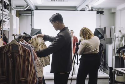 Side view of male designer choosing clothes from rack during photo shoot in studio