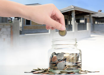 Cropped hand putting coin in jar on table against building