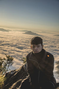 Portrait of young man sitting on mountain against sky