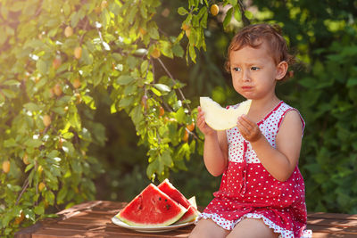 Little curly white girl eating melon in the garden , green blurred background