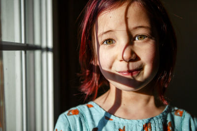 A cute little girl with dyed red hair stands in patch of window light