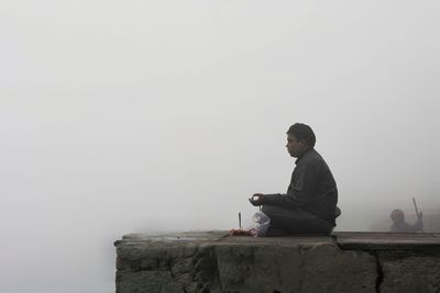 Side view of young man meditating against sky during foggy weather