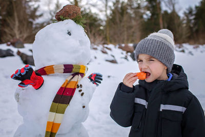 Boy in hat bites carrot, which was the nose of snowman, smiling. winter family weekend, lifestyle