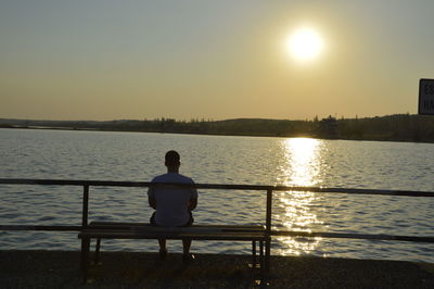 Rear view of man sitting on shore against sunset sky