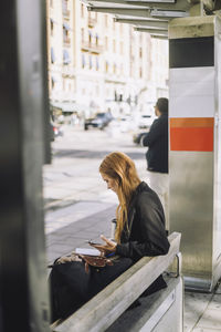 Side view of female freelancer sitting with smart phone at bus stop