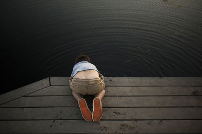 High angle view of boy looking in lake while kneeling on pier at park