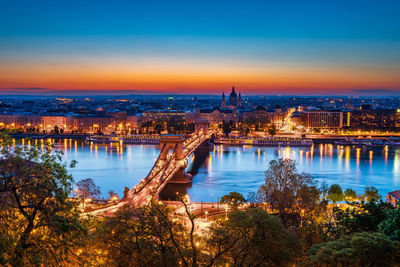 Aerial view of illuminated city at riverbank during sunset