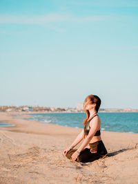 Side view of young woman doing yoga at beach against sky
