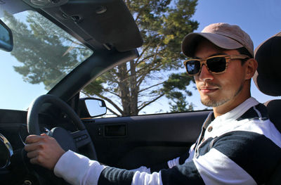 Young man with wooden sunglasses and baseball cap driving a convertible