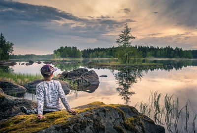 Rear view of boy sitting by lake against sky