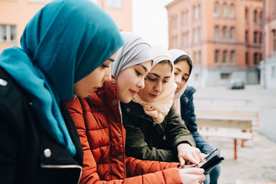 Female friends wearing hijabs sitting in city sharing digital tablet