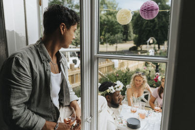 Young man sitting at window while looking at friends enjoying dinner party outside cafe