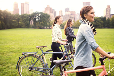 Women with bicycles walking on field in park against city