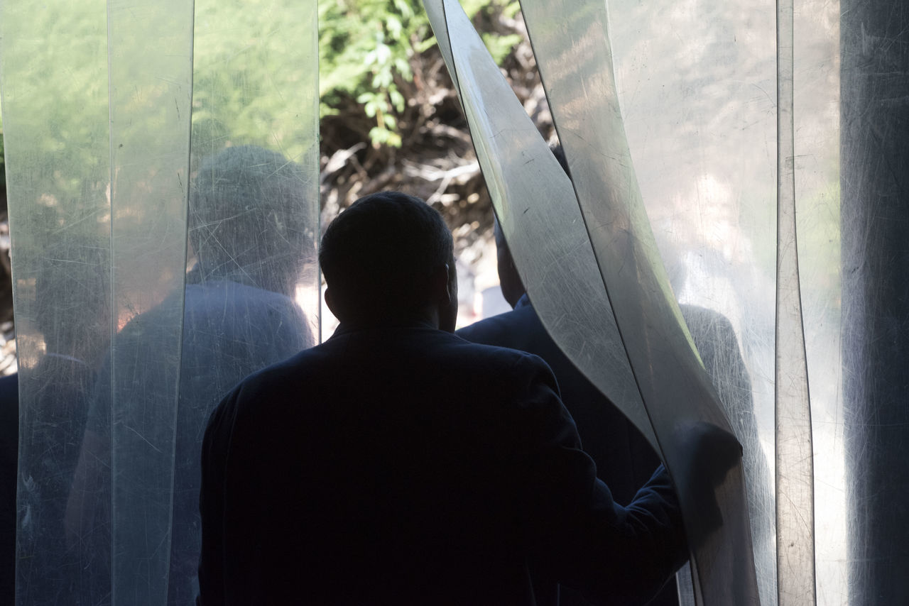 REAR VIEW OF MAN LOOKING THROUGH WINDOW IN GLASS