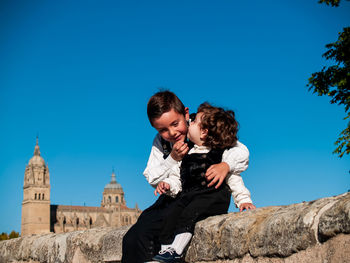 Cute boy kissing brother sitting on wall against historic building