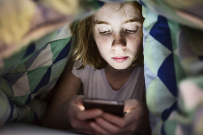 Girl lying under blanket in bed looking at her smartphone