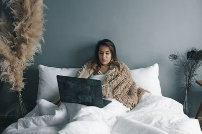 Sick young woman using laptop on bed against wall at home