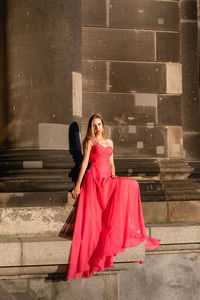 Full length of young woman in pink evening gown sitting against columns