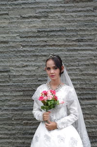 Portrait of bride holding bouquet while standing against wall